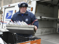 A man with a white beard in a blue postal uniform loads a tray of letters in to the back of a postal truck.