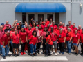 Forty workers in red t-shirts pose together outside a building with a blue awning.