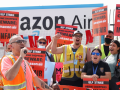 Workers in orange vests yell together, outdoors in front of an Amazon Air sign. Person in front has a microphone. Several have red printed picket signs saying "ULP strike. Prime shoppers beware: Amazon Air is unfair." Or the same in Spanish.