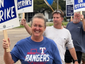 A woman with a blue Texas Rangers T-shirt and a sign that says “On Strike” smiles as she leads a line of autoworkers towards the camera.