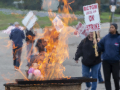 Strikers march holding picket signs that read "On Strike," while a fire rises from a burn barrel to keep them warm.