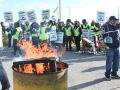 In the foreground a burn barrel, one striker walking purposefully, one raising a fist. In the background a large crowd of strikers carrying "CUPW on strike" picket signs and wearing safety-yellow vests.