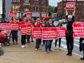 About 15 people, mostly white women, stand on a street corner wearing big red printed signs that say "Haverill educators on strike!" One woman holds her sign high in the air and turns to face the others, in profile to the camera, with a lively open-mouthed expression, maybe yelling or singing. Her sign says "Fair contract now." 