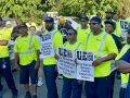 A group of sanitation workers, most in their bright green hi-visibility work vests, hold signs saying, "Fair Pay for City Workers!" and "City Workers Can't Afford to Live in the City we Serve!" during a rally.