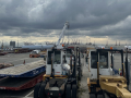 Small port vehicles are in the foreground, and harbor cranes are visible in the distance.