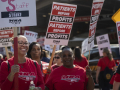 a group of smiling nurses, mostly African American, in red shirts hold signs saying ‘patients before profits’