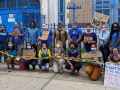 21 adults in masks outside a school. Together they hold "CAUTION" tape. Handmade signs say "100% remote start," "no mas muertes," "ready or not," "we demand safe schools," "no more martyrs for the DOE #Remote until it's safe" and "people got sick in this buidling."