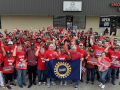 A group of a hundreds autoworkers gather outside with red shirts and fists raised.