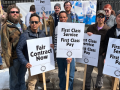 A dozen people, mostly men, face the camera resolutely on a sunny cold day outside a post office. Several are holding printed signs that say "Fair contract now" or "First class service, first class pay" with the NALC Branch 9 logo.