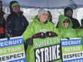 Six smiling people with green and blue strike signs stand under a tent in the snow.