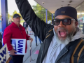 A Black man in sunglasses with an exuberant expression exits a factory turnstile with his right arm raised.