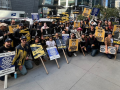 A large, upbeat crowd of picketers poses on a midtown Manhattan sidewalk, many kneeling. Some picket signs say "SAG-AFTRA on strike" and others say "IATSE supports workers' rights" or "IATSE Local 52 supporters workers' rights."