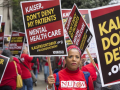 Strikers in red march with printed picket signs. In the center is a Black woman with a serious expression. Her sign reads "Kaiser, don't deny my patients mental health care. KaiserDontDeny.org."