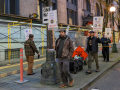 Four workers carrying picket signs walk in a circle on a downtown corner in dawn or twilight. One makes eye contact with camera.