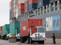 Four port workers in hardhats and neon vests look tiny as they stand beside a line of trucks with a stack of huge containers behind them.