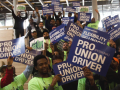 A big crowd of workers in matching green T-shirts hold up printed signs reading "PRO(FESSIONAL) UNION DRIVER"