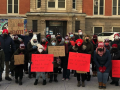 25 people in front of a school, masked and bundled up. Handmade signs read: "Keep our parents, teachers, students safe!""Safe schools should not be a debate!" "Let us teach!" "15 out of 18 classes in my school had at least 1 covid positive case" "Porque tenemos que suplicar para tener escuelas seguras?" (in Spanish: Why do we have to beg to have safe schools?) "Little Village has a positivity rate of 26.9%" "Lori let us teach (remotely) until Jan. 18th #EndTheLoriLockout"