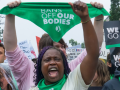 Women shout and raise their fists at a rally. Many are wearing green bandannas and one is holding the bandanna up in the air: it says "Bans off our bodies." A sign held up in background says "We won't go back."