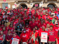 Large group of people outdoors, most wearing red shirts, some holding picket signs, on a sunny day. They are posed for a group photo on the steps of a school. The photo shows more than 200 people and it looks like there are more outside the frame.