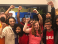 An exuberant group of young workers, diverse in race and gender, pose in a huddle and throw fists in the air