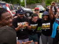 A young, racially diverse group of workers in "Amazon Strike" T-shirts huddles in a parking lot together, cheering. Some raise fists in the air. Behind them a Teamsters Local 705 truck is visible.