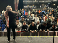 A large crowd of workers in a dark auditorium listen to a worker onstage. The speaker's back is to us and she wears a head scarf or hijab. Some in the audience are smiling, some look focused, and two in the front row are animatedly discussing something. The workers are racially diverse, including many who could be of South Asian or East African descent, as the article describes.