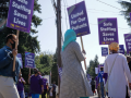 Workers picket in a circle. Many carry purple printed SEIUHealthcare signs that say "Safe Staffing Saves Lives" or "United for Our Patients." In the foreground is one worker viewed in profile and three silhouetted from behind; two of these workers wear head scarves and one wears the conical style of hat common in Southeast Asia.