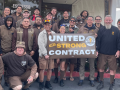 26 people (mostly men) in brown UPS uniforms pose outside Duke's coffee shop, smiling. Some hold a banner: "United for a strong contract" with the UPS Teamsters United logo.