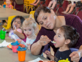 A woman spoon-feeds a toddler seated at a table next to two more toddlers, also eating. Visible behind her is another row of kids in high chairs.