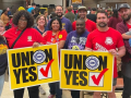 Ten smiling people pose together in a crowded meeting room. Two are holding huge yellow "Union Yes" signs with the UAW logo. Most are wearing red, blue, or purple T-shirts with UAW logo or organizing-themed slogans. The man on the far right holds a small child in his arms.