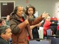 A person holding a mic stands speaking, gesturing with one arm, while others in the meeting listen intently. The group is diverse in race, gender, and age.