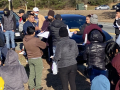 A group of workers stand by the side of the road while and man and woman pass out information to them.