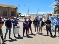 A group of workers, some holding drums, gathers in a circle outside a port.