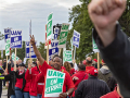 GM workers on a picket line, one work offscreen with arm raised, another centered with sign with arm raised.