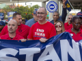 A group of men and women in red T-shirts stand holding a blue banner, the word ‘Stand’ is visible.