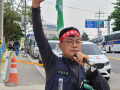 A man, Yang Hoe-dong, walks along a sidewalk wearing a resolute expression. His right arm is in the air, holding a green flag. In his left hand he holds a microphone to his lips. He wears a red headbank with printing on it in Korean, and a vest and badge that also have printed text in Korean. It seems likely he is speaking or leading chants at a union protest.