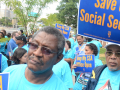 A crowd dressed in light blue T-shirts holds signs saying Save our Social Security and Keep My SSA Office Open.