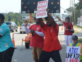 Black and white men, most in red union T-shirts, picket energetically on a streetcorner. Man in front, in turquoise shirt, is talking or chanting into a mic, arm outstretched. Everyone's pose is active. Printed signs say "honk if you support workers" and "AT&T unfair, CWA on strike, ULP." One handmade sign, partially visible, says "AT&T Unfair Labor." The man in the center is holding his sign high and wind or movement blows his red shirt.