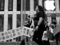 People march in front of an Apple store in New York City, holding signs and banners in English and Chinese. Their faces are blurred out in the photo due to fears of retaliation from the Chinese government against them and their families.