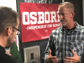 A man in a checkered shirt talks to two people with their backs to the camera in front of a red “Dan Osborne Independent for Senate” banner.