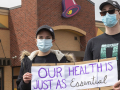 Two taco bell workers with masks holding up a sign in front of a taco bell that says "our health is just as essential."