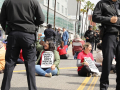 Several workers sit in the street with signs saying “Kaiser Don’t Deny Mental Health”--they are surrounded by police.