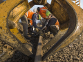 Workers in orange vests and hardhats crouch around a track. They are framed by a big yellow piece of equipment in the foreground. The day is very sunny.