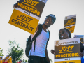 A young slender Black man in a baseball cap smiles down at the camera with a sign saying “Just Practicing for a Just Contract.” To the left of him is a Black woman in sunglasses with a whistle in her mouth carrying the same sign..