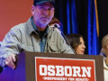 A man in a ball cap stands behind a lectern that says Osborn for Senate