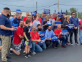 A group of workers in red, blue, or grey shirts hold signs saying “United for a Strong Contract,” and “COLA and Fair Pay Now”  