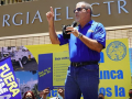 Man in blue shirt with right arm raised and left arm holding microphone addressing crowd, Fuera LUMA flag visible in foreground, building of Puerto Rico Electric Power Authority (PREPA)