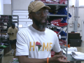 A Black man in a baseball cap and a T-shirt that says "Home" with the shape of Africa for the "O" speaks to the camera. He is standing in a warehouse, in front of shelves stacked high with folded apparel . Another Black man can be seen behind him, working.