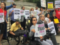 Demonstrators, some in wheelchairs and some not, stand in front of a public building. Many are holding signs: "ACCESSIBLE TRANSIT FOR ALL," "ELEVATORS NOW," "STRANDED BY CUOMO" 