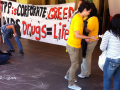 Activists draw chaik outlines on the ground in front of a hotel entrance; one outline is labeled "Vietnam." Others hold a banner: "TPP is corporate greed. AiDS drugs = life." 